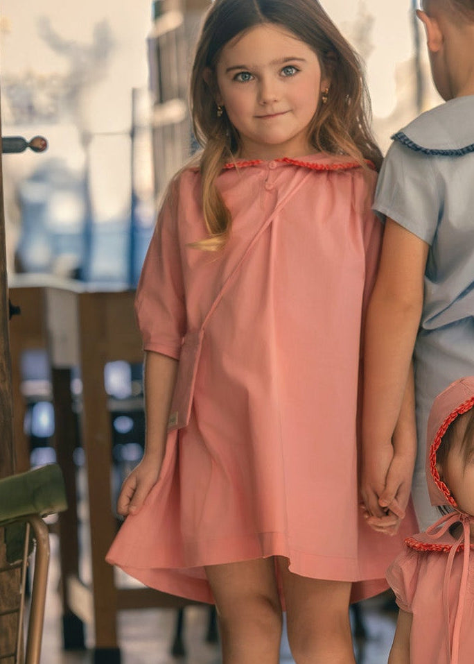 Three children in dresses standing in a room with large windows.